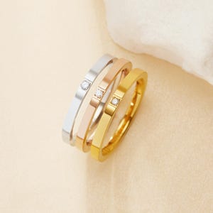 May include: Three stackable rings in silver, rose gold, and gold. Each ring features a small, clear gemstone. The rings are simple, modern, and suitable for everyday wear. The rings are made of a shiny metal.