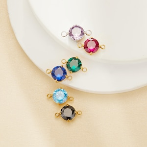 May include: A collection of colorful gemstone connectors. The gemstones are round and come in various colors, including purple, red, green, blue, light blue, and black. Each gemstone is set in a gold-colored metal setting with small loops on each side.