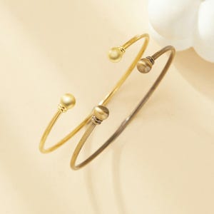 May include: Two gold bangle bracelets with round ball ends. One bracelet is a lighter gold color and the other is a darker gold color.