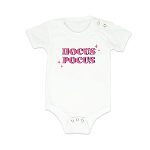 Può includere: Body bianco per neonato con testo rosa che recita "Hocus Pocus" con stelle rosa su entrambi i lati del testo.