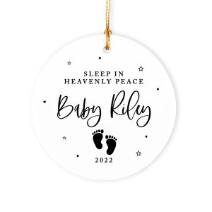 May include: White ceramic ornament with black text that reads "Sleep in Heavenly Peace Baby Riley" and a pair of black footprints. The year 2022 is printed below the footprints.