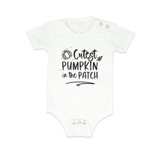 Può includere: Body da bambino bianco a maniche corte. Il body presenta la scritta "Cutest PUMPKIN in the PATCH" in nero, con una grafica di zucca e foglia. Ha chiusure a scatto sul fondo e sulla spalla.