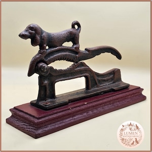 May include: Vintage cast iron dachshund nutcracker. The dog figure sits on the lever mechanism. The nutcracker is mounted on a stained, dark red wooden base. The metal has a dark brown, aged finish.