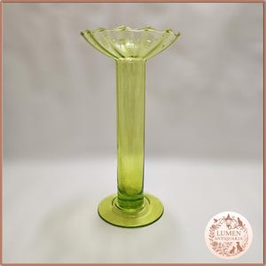 Peut inclure: Un vase en verre vert clair avec un dessus évasé et festonné et une base circulaire. Le vase a une longue tige cylindrique. Le verre a une légère teinte jaunâtre. Le vase est probablement à des fins décoratives.