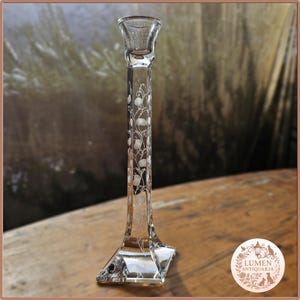 Pu&ograve; includere: Un candeliere in vetro trasparente con una base svasata e un sottile stelo inciso. Lo stelo presenta un delicato motivo floreale. Il candeliere &egrave; esposto su una superficie di legno.