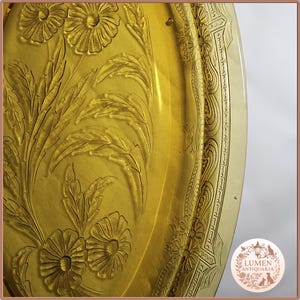 Op de afbeelding: Close-up van een goudgeel antiek bord met reliëf bloemen- en bladmotieven. Het bord heeft een gedetailleerde rand met ingewikkelde patronen en tekst. Het bord heeft een licht doorschijnend uiterlijk.