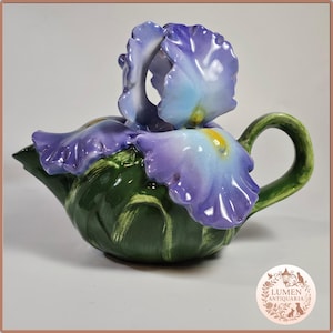 Könnte beinhalten: Eine Keramik-Teekanne in Form einer Irisblüte. Die Teekanne hat einen grünen Boden und Griff, mit lila und blauen Blütenblättern, die den Deckel bilden. Der Ausguss befindet sich links und der Griff rechts.