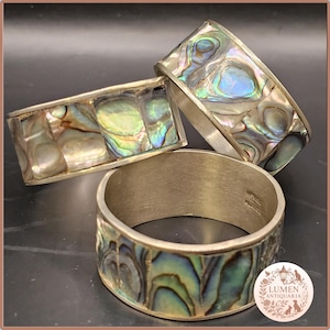 Könnte beinhalten: Drei silberne Serviettenringe mit irisierenden Abalone-Muscheleinlagen. Die Abalone-Muschel zeigt eine Reihe von Farben, darunter Blau, Grün und Lila. Die Serviettenringe werden auf einer dunklen Oberfläche präsentiert.