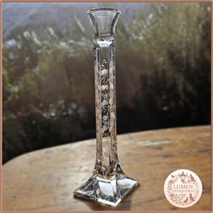 Pu&ograve; includere: Un candelabro di vetro trasparente con base quadrata e stelo alto e sottile. Lo stelo &egrave; decorato con un motivo floreale inciso. Il candelabro &egrave; progettato per contenere una singola candela.