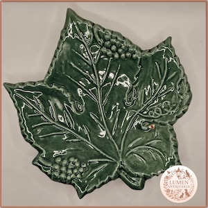 Puede incluir: Un plato verde de cerámica con forma de hoja y acabado brillante. El plato presenta vetas detalladas y un racimo de uvas, con un borde decorativo. El diseño recuerda a una hoja de vid.