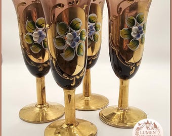 May include: Four ornate champagne flutes with gold stems and bases. The glasses have a dark brown lower section, a gold center with floral designs, and a pink upper section. The floral design includes green leaves and purple and white flowers.