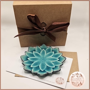 Puede incluir: Un plato de cerámica turquesa con forma de flor de loto con una caja de regalo marrón atada con una cinta marrón. La caja es de cartón. Una tarjeta con texto y un logotipo está al lado del plato.