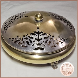 May include: An antique silver and gold-toned metal container with a decorative pierced lid. The lid features intricate floral cutouts and a small, round knob. The container has a rounded base and rests on three small feet.