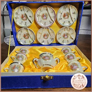 Może przedstawiać: Zestaw do herbaty z porcelany w stylu vintage w niebieskim pudełku. Zestaw zawiera sześć spodków, sześć filiżanek i cukiernicę. Każdy element jest ozdobiony kolorowym wzorem kwiatowym i figuralnym, ze złotym wykończeniem. Zestaw jest osadzony w żółtej satynowej wyściółce.