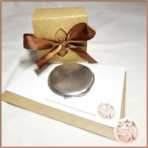 May include: A silver compact mirror with a textured design sits on a white card, next to a small brown gift box tied with a brown satin ribbon. The card has text and a circular logo.
