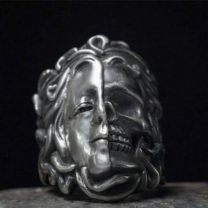 May include: A silver ring with a detailed design of a woman's face on one side and a skull on the other. The ring is made of metal and has a textured surface.
