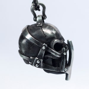 May include: A silver skull pendant with a detailed, textured surface. The skull is adorned with a metal cage-like structure, featuring a pointed, curved piece extending from the front.