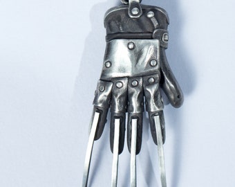 May include: A silver necklace with a pendant shaped like a glove with sharp claws. The glove is detailed with rivets and a silver band around the wrist.