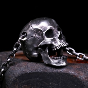 Può includere: Un ciondolo a forma di teschio in argento con un design dettagliato, appeso a una catena in argento. Il teschio ha la bocca aperta ed è posizionato su uno sfondo scuro.