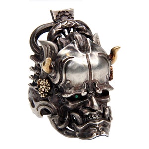Puede incluir: Un anillo de plata con un dise&ntilde;o detallado de una m&aacute;scara de demonio japon&eacute;s. La m&aacute;scara tiene ojos verdes, cuernos dorados y tallas intrincadas.