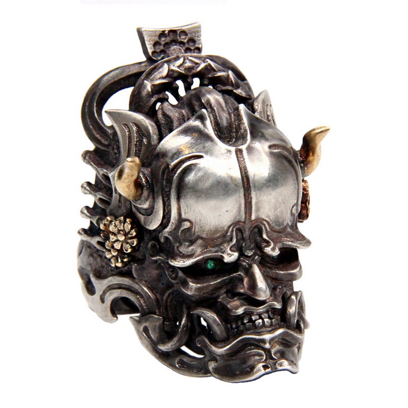Puede incluir: Un anillo de plata con un dise&ntilde;o detallado de una m&aacute;scara de demonio japon&eacute;s. La m&aacute;scara tiene ojos verdes, cuernos dorados y tallas intrincadas.