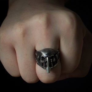 May include: A silver ring with a detailed skull design. The ring is worn on a finger and the skull is facing forward.