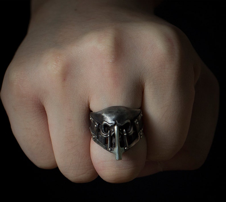 May include: A silver ring with a detailed skull design. The ring is worn on a finger and the skull is facing forward.