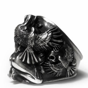 May include: A silver ring with a detailed design of two birds and a skull. The birds are in flight and the skull is in the center of the ring.