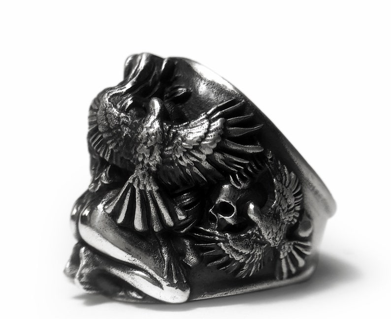 May include: A silver ring with a detailed design of two birds and a skull. The birds are in flight and the skull is in the center of the ring.