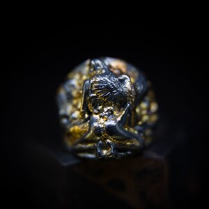 May include: A silver and gold ring with a detailed design of a figure in a seated position. The figure is surrounded by intricate details and small red gemstones.