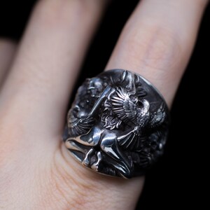 May include: A silver ring with a detailed design of a winged figure and a bird. The ring is worn on a finger.