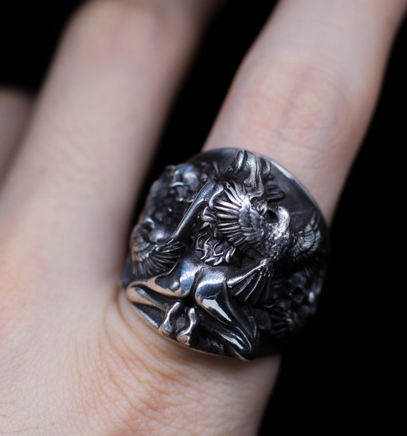 May include: A silver ring with a detailed design of a winged figure and a bird. The ring is worn on a finger.