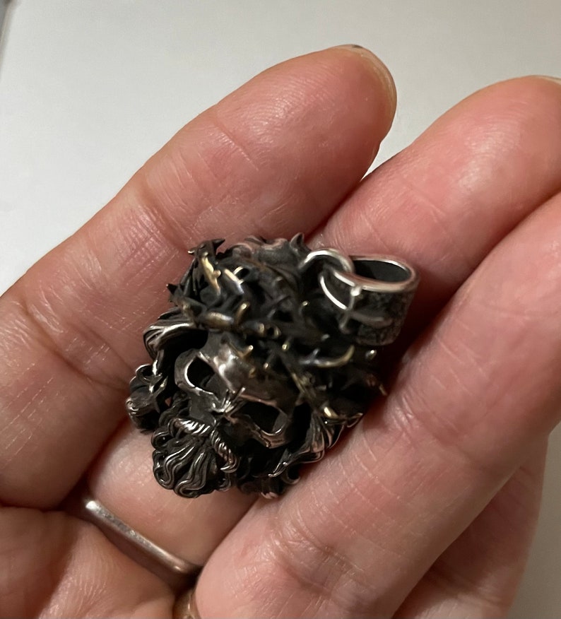 May include: A silver skull pendant with a crown of thorns and a beard. The pendant is detailed and intricate.