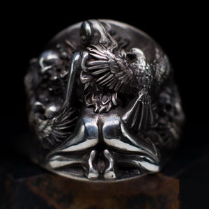 May include: A silver ring with a detailed design of a woman with wings, possibly an angel, holding a bird in her arms. The ring is made of metal and has a textured surface.