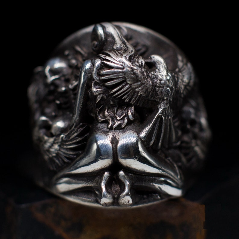 May include: A silver ring with a detailed design of a woman with wings, possibly an angel, holding a bird in her arms. The ring is made of metal and has a textured surface.