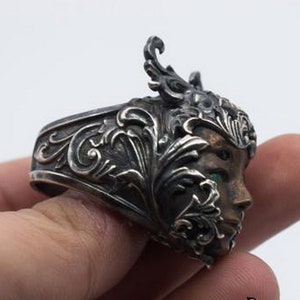 May include: A silver ring with an ornate design featuring a woman's face with green eyes. The ring is detailed with intricate floral patterns.