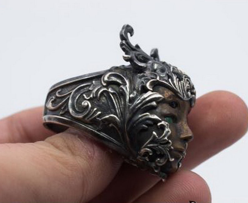 May include: A silver ring with an ornate design featuring a woman's face with green eyes. The ring is detailed with intricate floral patterns.