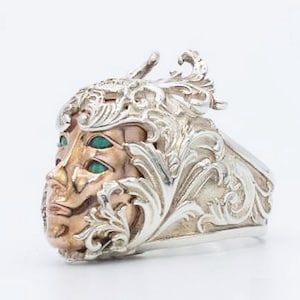 May include: A silver ring with an ornate design featuring a face with three green eyes. The face is surrounded by intricate floral details.