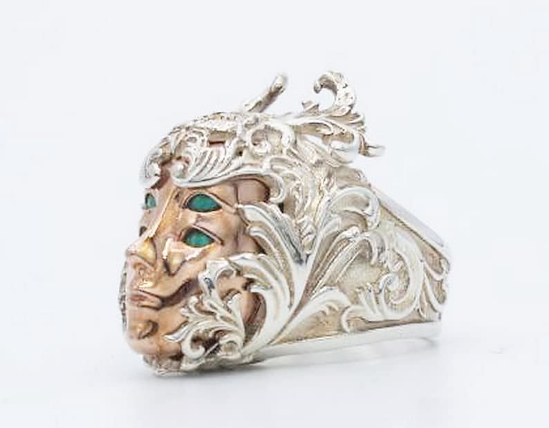 May include: A silver ring with an ornate design featuring a face with three green eyes. The face is surrounded by intricate floral details.