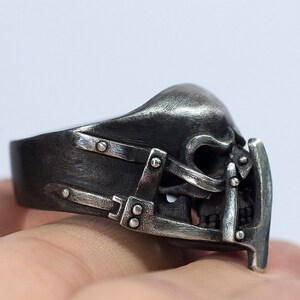 May include: A silver skull ring with a detailed design of a skull with a metal cage over the face. The ring is worn on a finger.