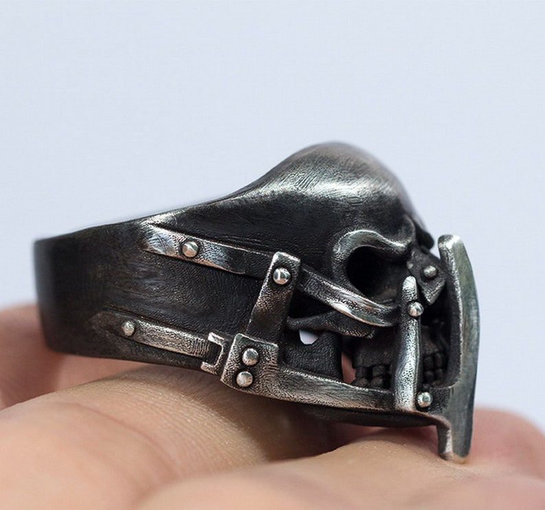 May include: A silver skull ring with a detailed design of a skull with a metal cage over the face. The ring is worn on a finger.