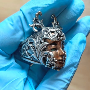 May include: A silver ring with an ornate design featuring a woman's face with green eyes. The face is made of copper and is surrounded by intricate silver floral details.