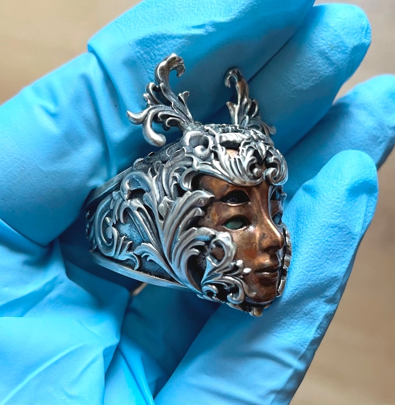 May include: A silver ring with an ornate design featuring a woman's face with green eyes. The face is made of copper and is surrounded by intricate silver floral details.