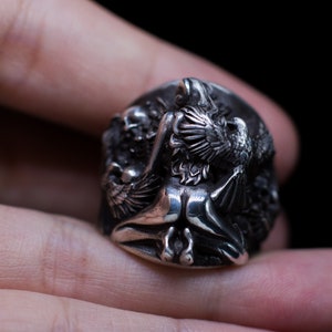 May include: A silver ring with a detailed design of a winged figure with a bird perched on its shoulder. The ring is worn on a finger.