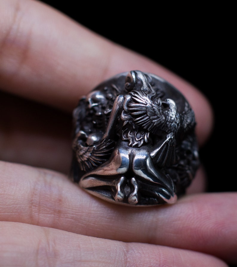May include: A silver ring with a detailed design of a winged figure with a bird perched on its shoulder. The ring is worn on a finger.