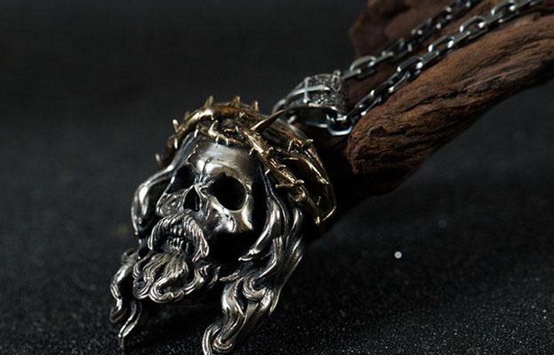 May include: A silver skull pendant with a gold crown of thorns, worn on a silver chain. The pendant is detailed with a beard and long hair.