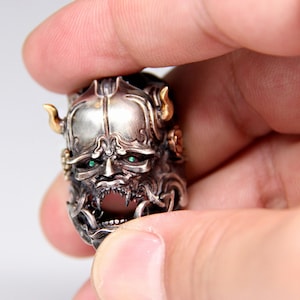 Puede incluir: Un anillo de plata con un dise&ntilde;o detallado de la cara de un demonio. El demonio tiene ojos verdes y una expresi&oacute;n feroz. El anillo est&aacute; hecho de plata y tiene un acento dorado.