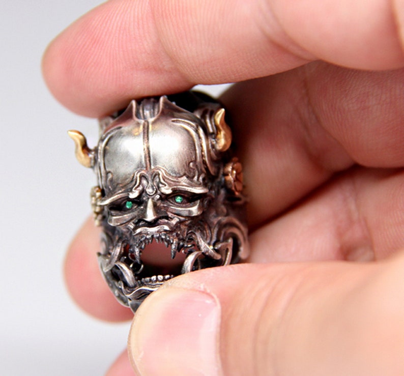 Puede incluir: Un anillo de plata con un dise&ntilde;o detallado de la cara de un demonio. El demonio tiene ojos verdes y una expresi&oacute;n feroz. El anillo est&aacute; hecho de plata y tiene un acento dorado.