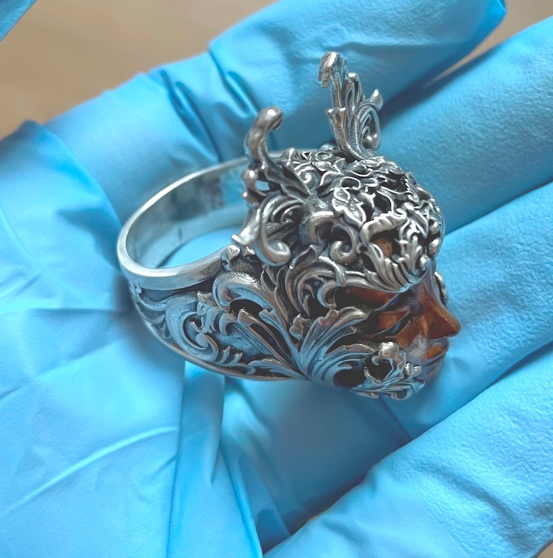 May include: A silver ring with a detailed design of a face with ornate floral accents.