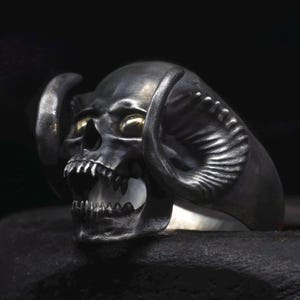 May include: A detailed, dark silver skull ring with curved horns and golden eye accents. The skull features a wide open mouth with visible teeth. The ring is set against a dark background, highlighting the intricate details of the design.